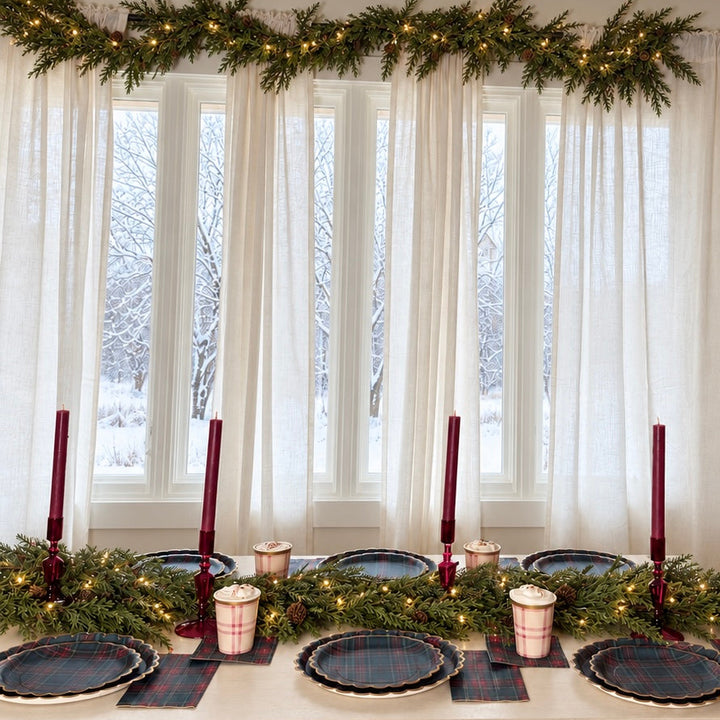 Christmas dinner table set with Heritage Tartan plates and napkins by snowy windows with garland and burgundy candles